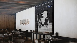 L’intérieur d’un restaurant présente des murs, des tables et des chaises en bois, ainsi qu’une grande fresque murale en noir et blanc.