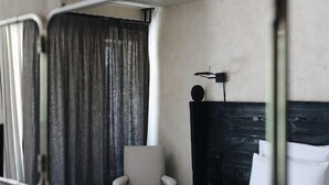 Un mur miroitant reflète une pièce dans laquelle on aperçoit un lit, une chaise et un rideau.