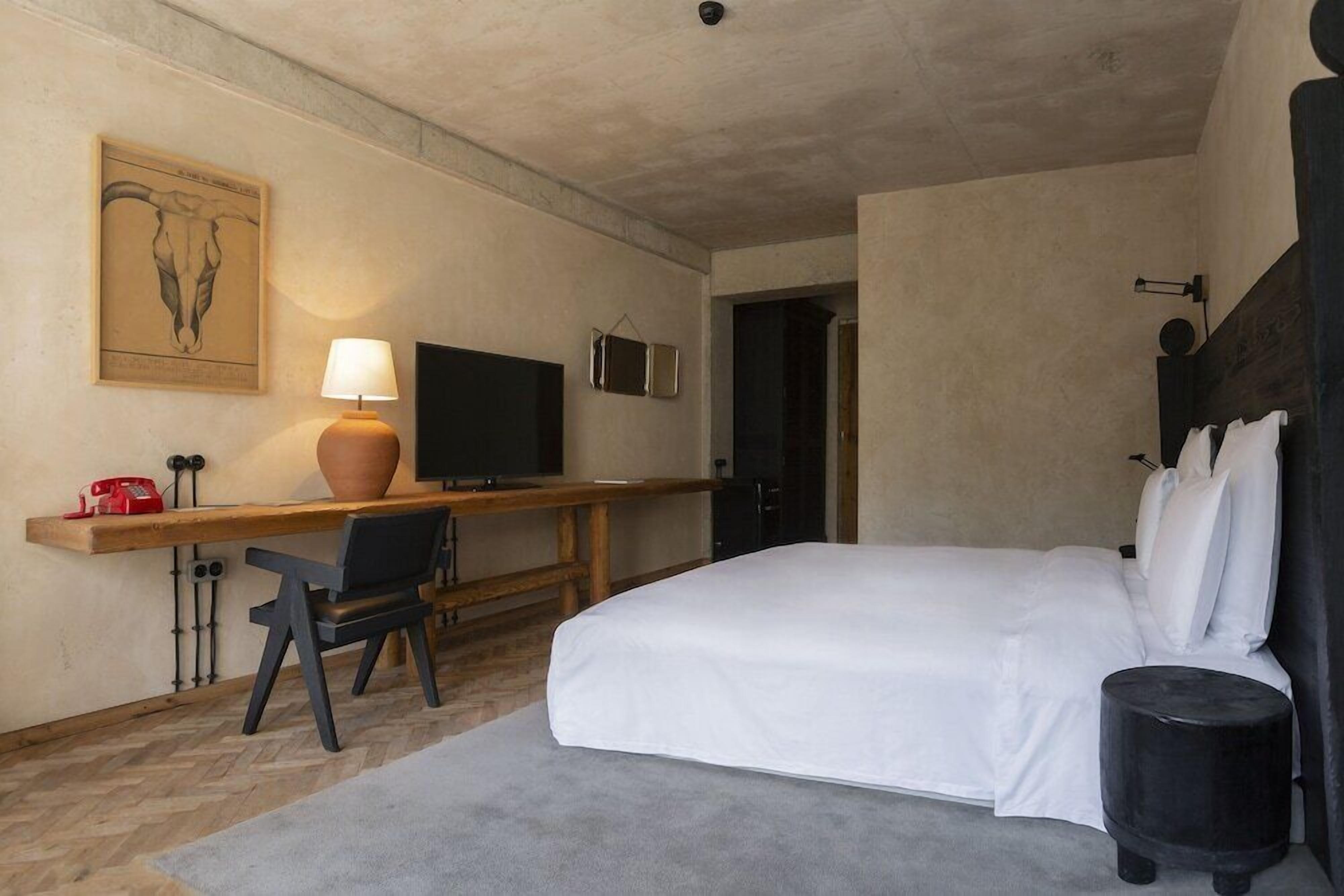 Et hotelværelse med seng, skrivebord med lampe, fjernsyn og et billede hængt på væggen.