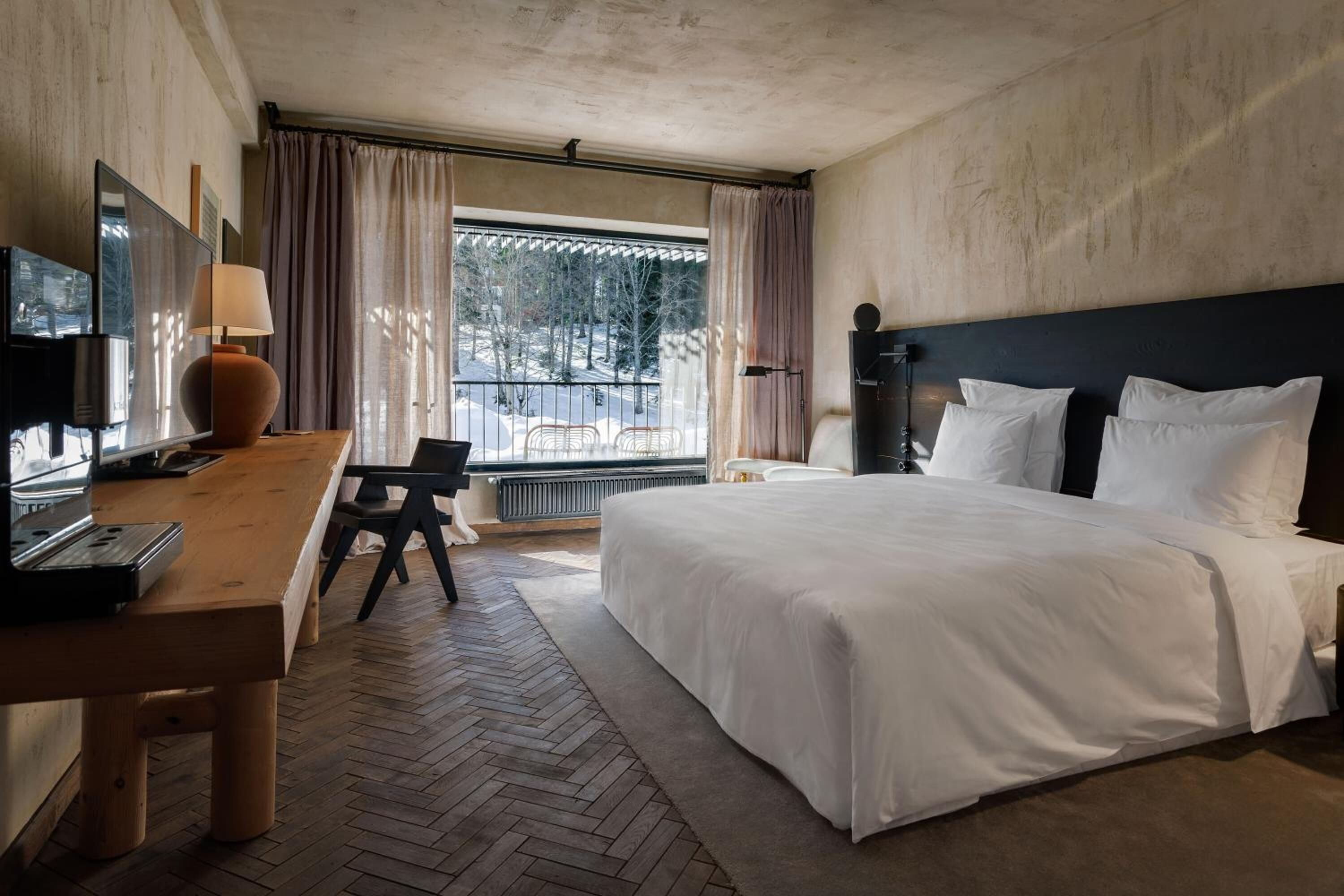 Ein Hotelzimmer mit einem großen Bett, einem Schreibtisch mit Lampe, einem Fernseher und Blick auf Bäume durch das Fenster.
