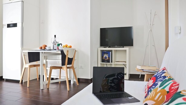 Uma sala de estar moderna com um laptop sobre uma mesa branca, uma TV em um suporte e uma almofada colorida.