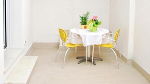 Um pequeno espaço de pátio fechado com uma mesa redonda e duas cadeiras amarelas, toalha de mesa branca e um vaso com flores.
