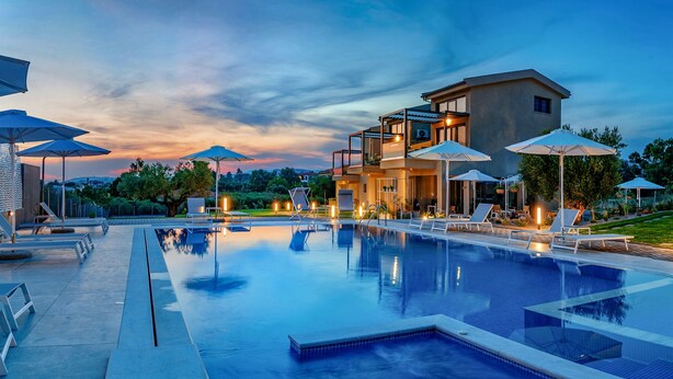 A swimming pool with lounge chairs and umbrellas, surrounded by modern buildings and a landscaped garden. - Miroes Suites (Sithonia)