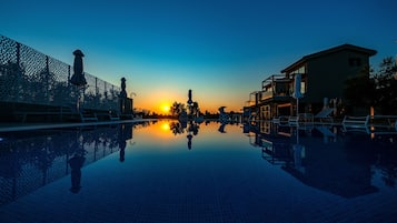 Un área de piscina tranquila con un reflejo claro del atardecer, una zona de estar cubierta y un edificio con balcones.
