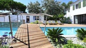 A wooden deck leading to a pool with lounge chairs and a white building in the background. - Spacious house with pool and beach access (Les Portes-en-Ré)