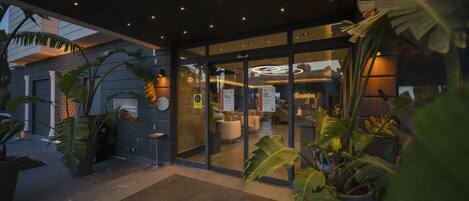 A modern hotel entrance with a reception area, glass doors, and potted plants.