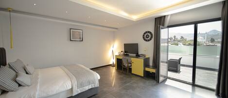 A modern hotel room with a bed, desk, and a view of the city through large windows.
