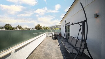 Una terraza en la azotea con barandilla de cristal, mobiliario exterior y vistas a la ciudad y las colinas.
