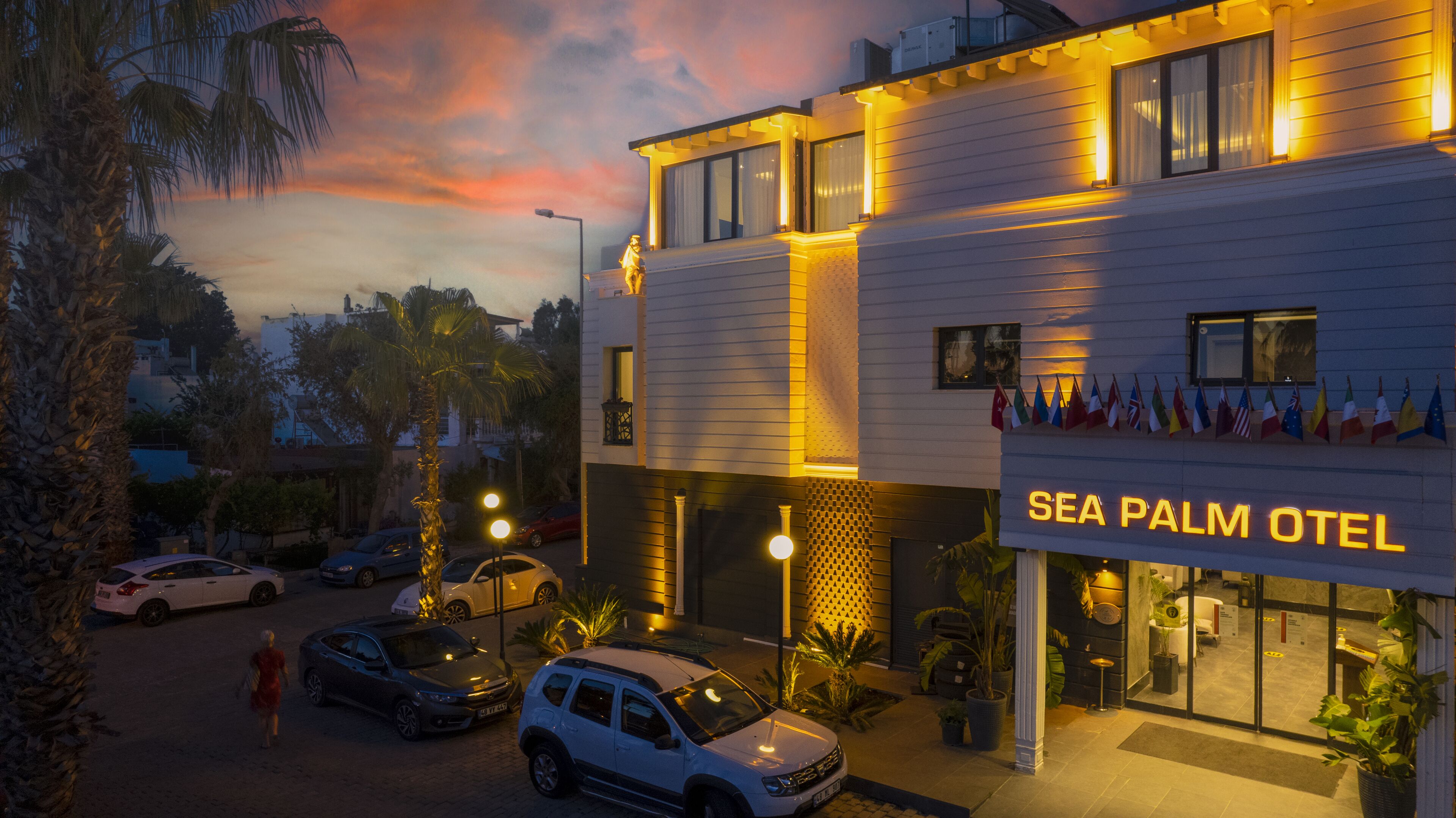 A hotel named "SEA PALM OTEL" with a modern design, featuring a prominent entrance, multiple flags, and a well-lit facade.