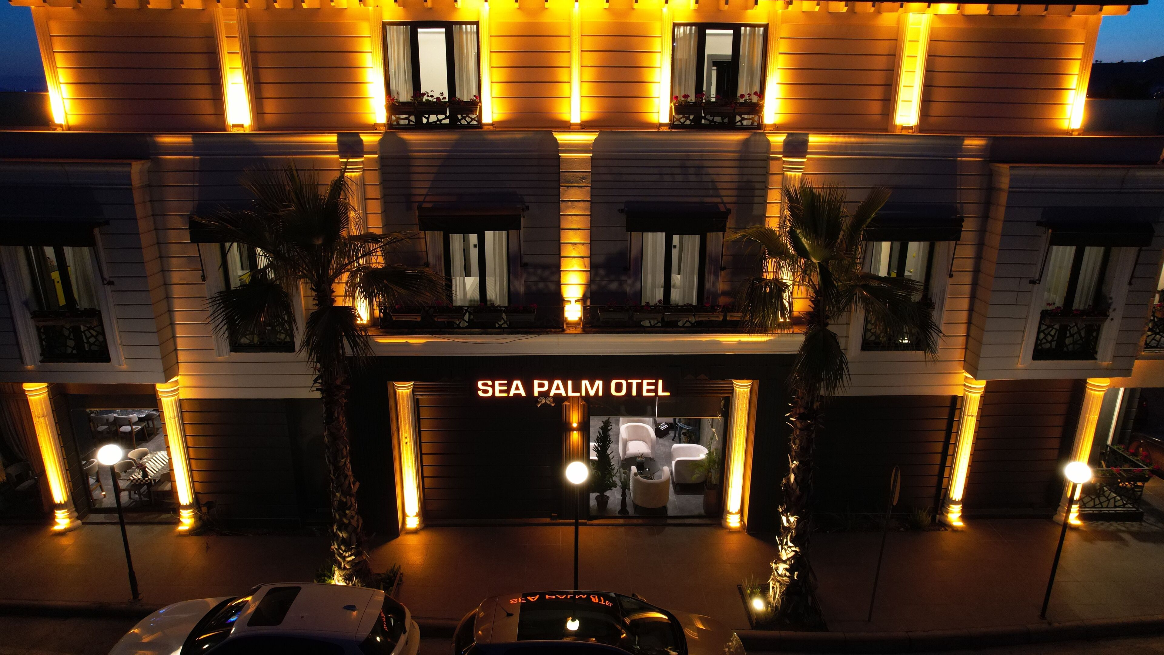 L’entrée d’un hôtel, avec une enseigne indiquant « SEA PALM OTEL », se distingue par sa façade moderne, ses fenêtres illuminées et son entrée centrale.