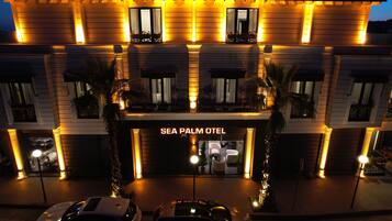 L’entrée d’un hôtel, avec une enseigne indiquant « SEA PALM OTEL », se distingue par sa façade moderne, ses fenêtres illuminées et son entrée centrale.