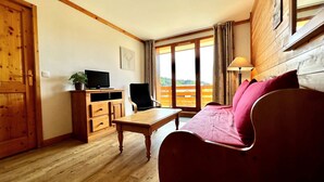 A room with a wooden floor, a bed with pink bedding, a wooden desk with a television, a chair, and a window with a view of a wooden deck and greenery.