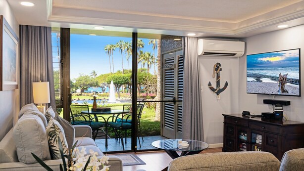 Habitación de hotel con balcón, sofá, televisor y vistas a palmeras y a la playa.
