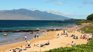 Una playa con gente tomando el sol y nadando, cielo despejado y montañas al fondo.