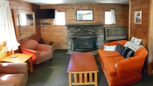 Ein gemütliches Blockhaus-Interieur mit Kamin aus Stein, Holzwänden, einer Couch, einem Sessel und einem kleinen Tisch.