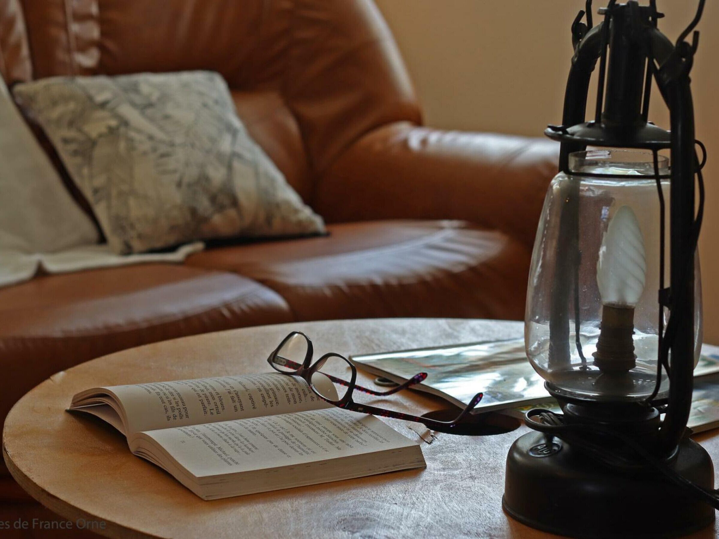 A leather sofa with a cushion, a round table with a book and glasses, and a black lantern.