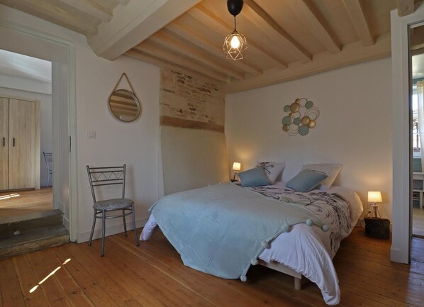 Un dormitorio con piso de madera, una cama con colcha azul claro, dos lámparas de noche, una silla y un candelabro.