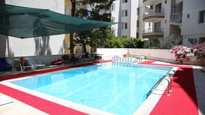 Una piscina con bordo rosso, zona d'ombra e vista su edifici condominiali sullo sfondo.