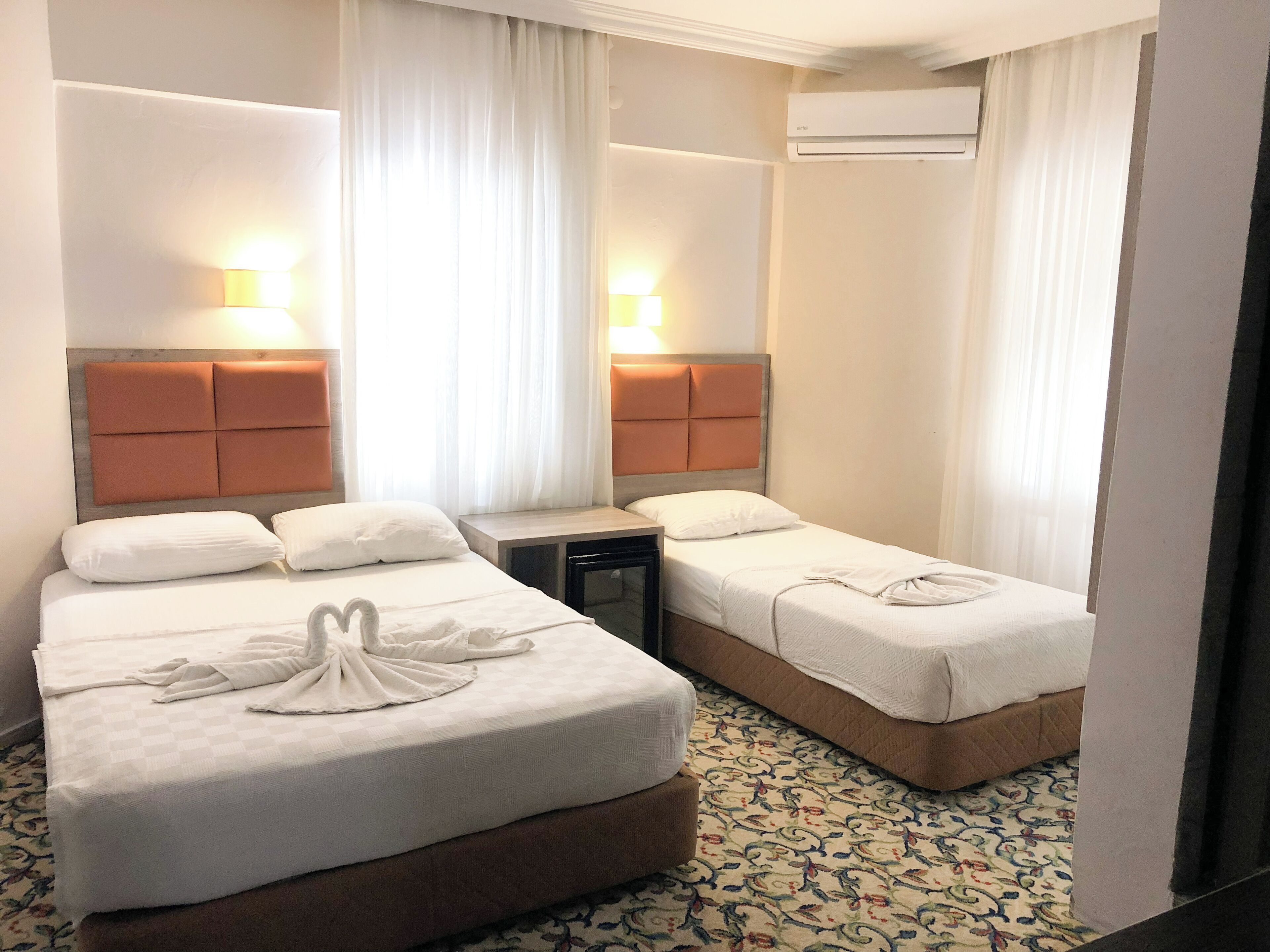 Habitación de hotel con dos camas, cada una con cabecera y lámparas de noche. Hay una mesita entre las camas, y un equipo de aire acondicionado montado en la pared.