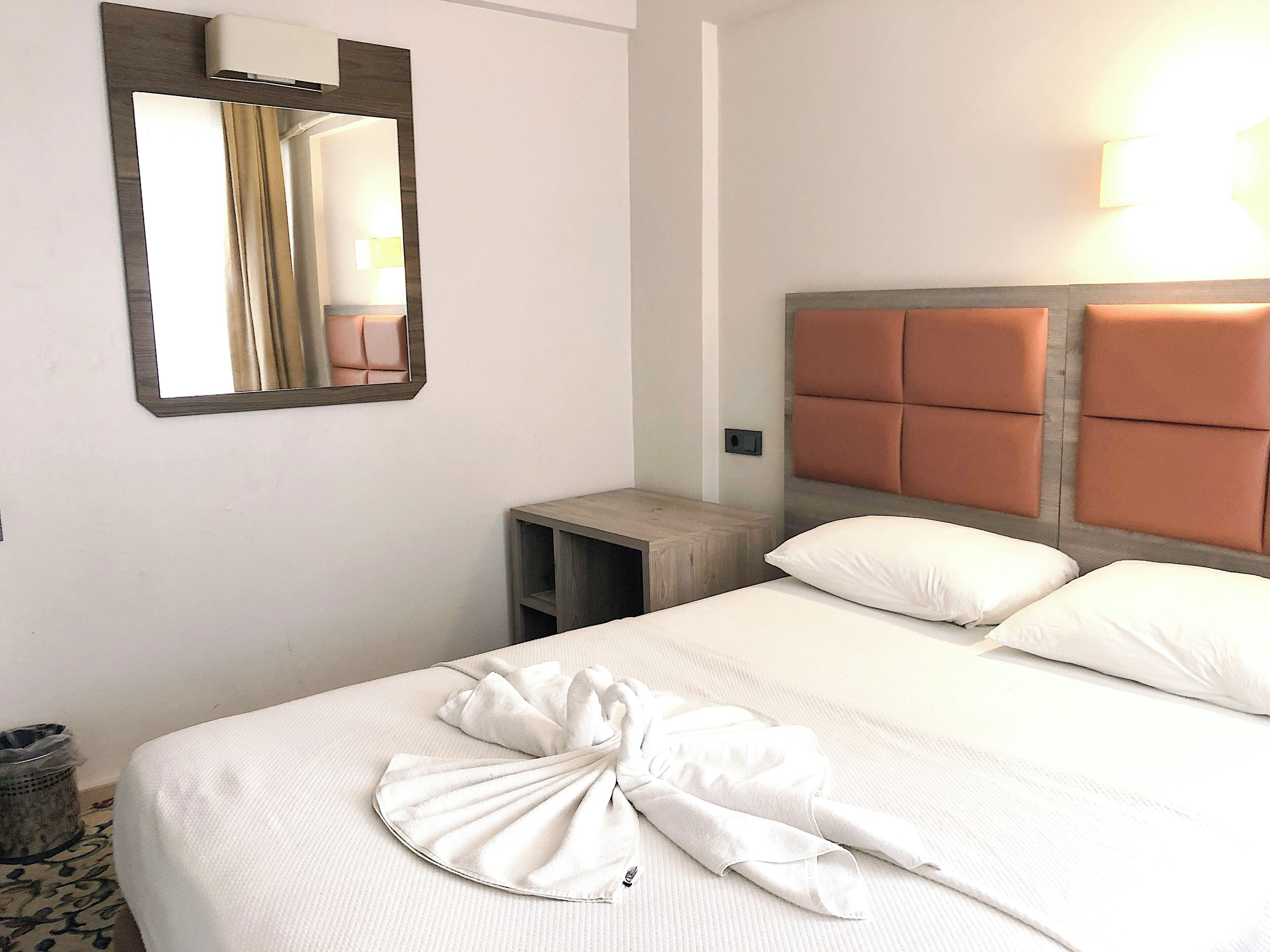 A hotel room with a bed, a bedside table, a mirror, and a wall-mounted light fixture.