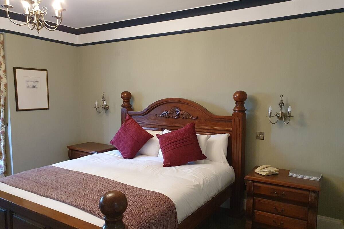 A neatly made bed with a wooden headboard, two red pillows, and a brown throw blanket. There is a nightstand with a telephone and a small wooden cabinet beside the bed.