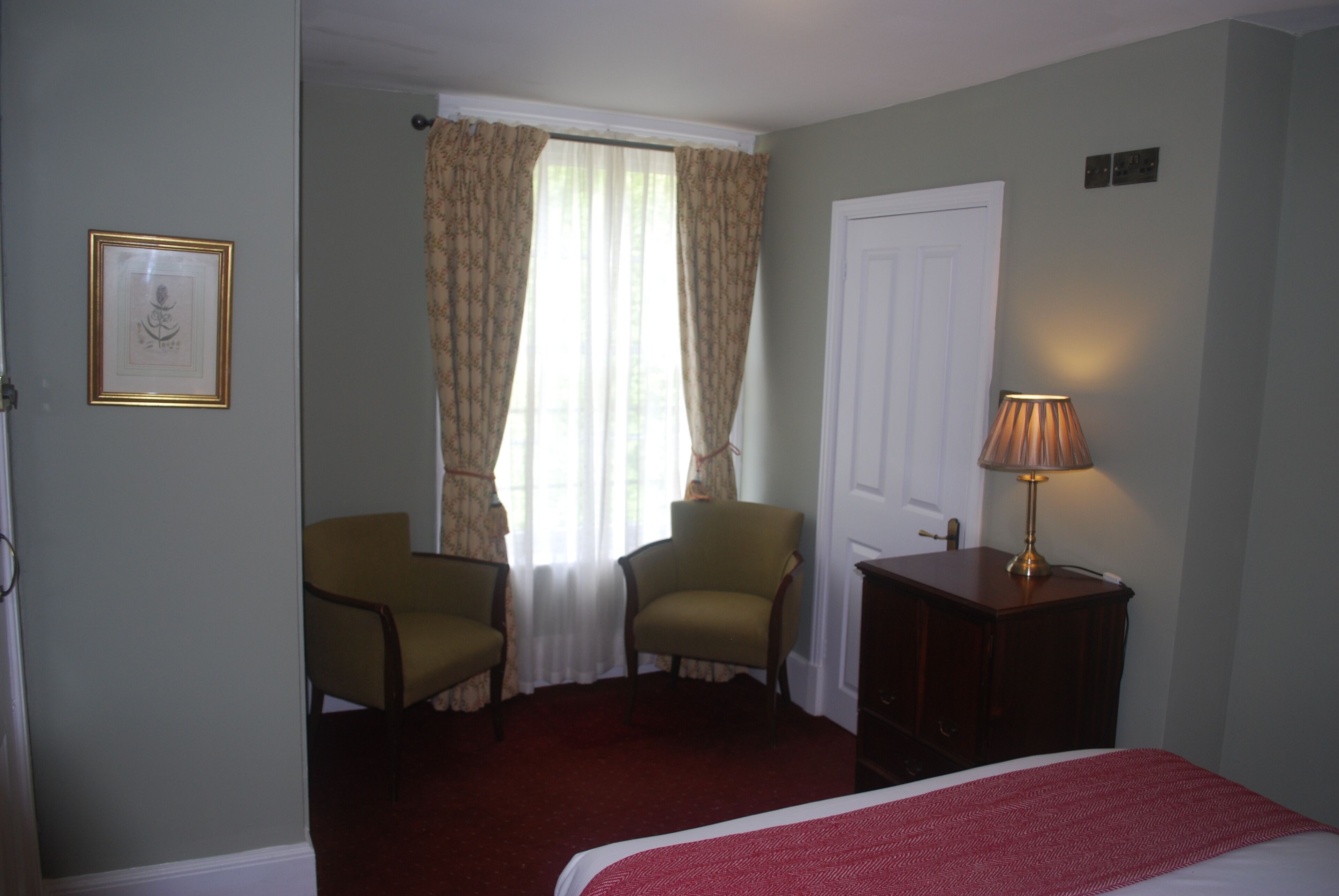 A hotel room with a bed, two armchairs, a wooden nightstand with a lamp, a window with curtains, and a framed picture on the wall.