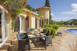 Un patio con una mesa dispuesta para dos, sillas de mimbre, una piscina pequeña y vistas a un paisaje montañoso.