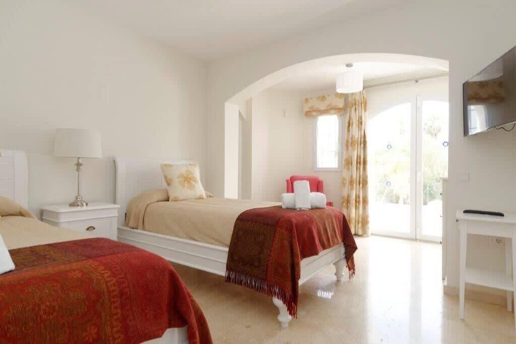 A hotel room with two beds, a TV, a white nightstand, a lamp, and a view of the outdoors.
