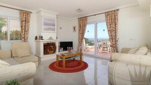 Un salon avec un téléviseur, un canapé et une table. Il y a un balcon avec des chaises et une vue sur l’extérieur.