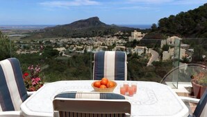 Une table avec un saladier de fruits et des verres, donnant sur un paysage pittoresque avec une colline et des bâtiments.
