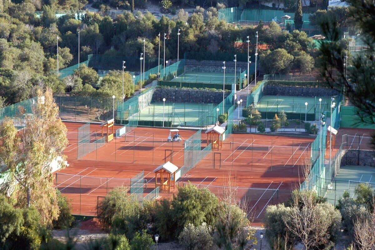 A tennis court complex with multiple courts, surrounded by trees and a green fence.