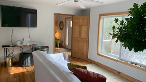 A modern living room with a white sofa, a ceiling fan, a TV mounted on the wall, and a large window with a view of the outdoors. - Exclusive walk to the beach Lazy Point House (Amagansett)