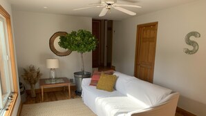 A cozy living room with a white sofa, a wooden floor, a ceiling fan, a potted tree, and a decorative letter 'S' on the wall. - Exclusive walk to the beach Lazy Point House (Amagansett)