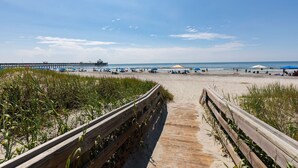 Una spiaggia con una passerella in legno che conduce alla riva, un molo sullo sfondo e ombrelloni.
