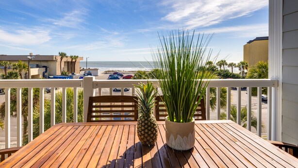 Un terrazzo in legno con tavolo, sedie, una pianta in vaso e un’ananas. Il terrazzo si affaccia su una spiaggia con palme e un’area parcheggio.
