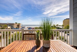 Une terrasse en bois avec une table, des chaises, un pot de fleurs et un ananas. La terrasse donne sur une plage bordée de palmiers et un espace de stationnement.
