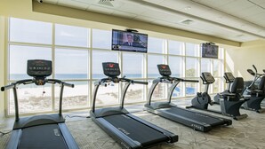Ein Fitnessstudio mit Laufbändern und Ergometer, großen Fenstern mit Blick auf den Strand und einem Fernsehbildschirm, der eine Nachrichtensendung zeigt.