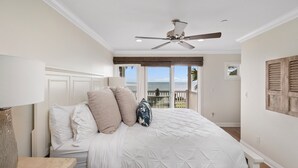 A bedroom with a bed, a ceiling fan, a nightstand, and a view of the ocean through large windows. - Sunset Dreams on the 101 (Carlsbad)