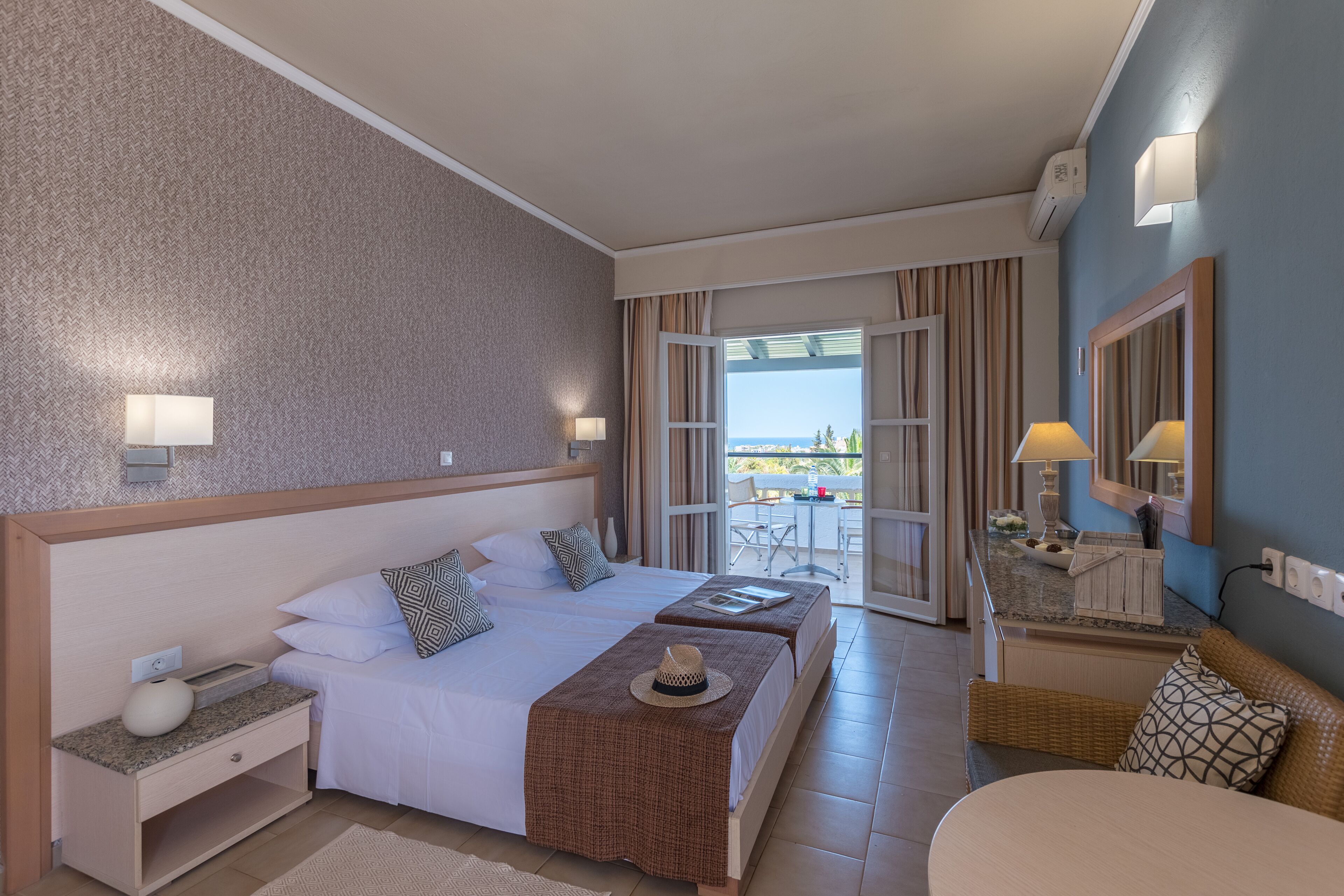 A hotel room with a large bed, a smaller bed, a desk, and a balcony with a table and chairs.