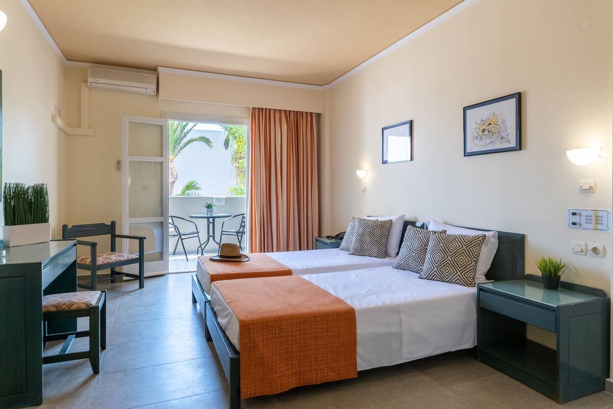 Habitación de hotel con dos camas, un escritorio, sillas, una mesita, una planta en maceta y vistas al exterior.