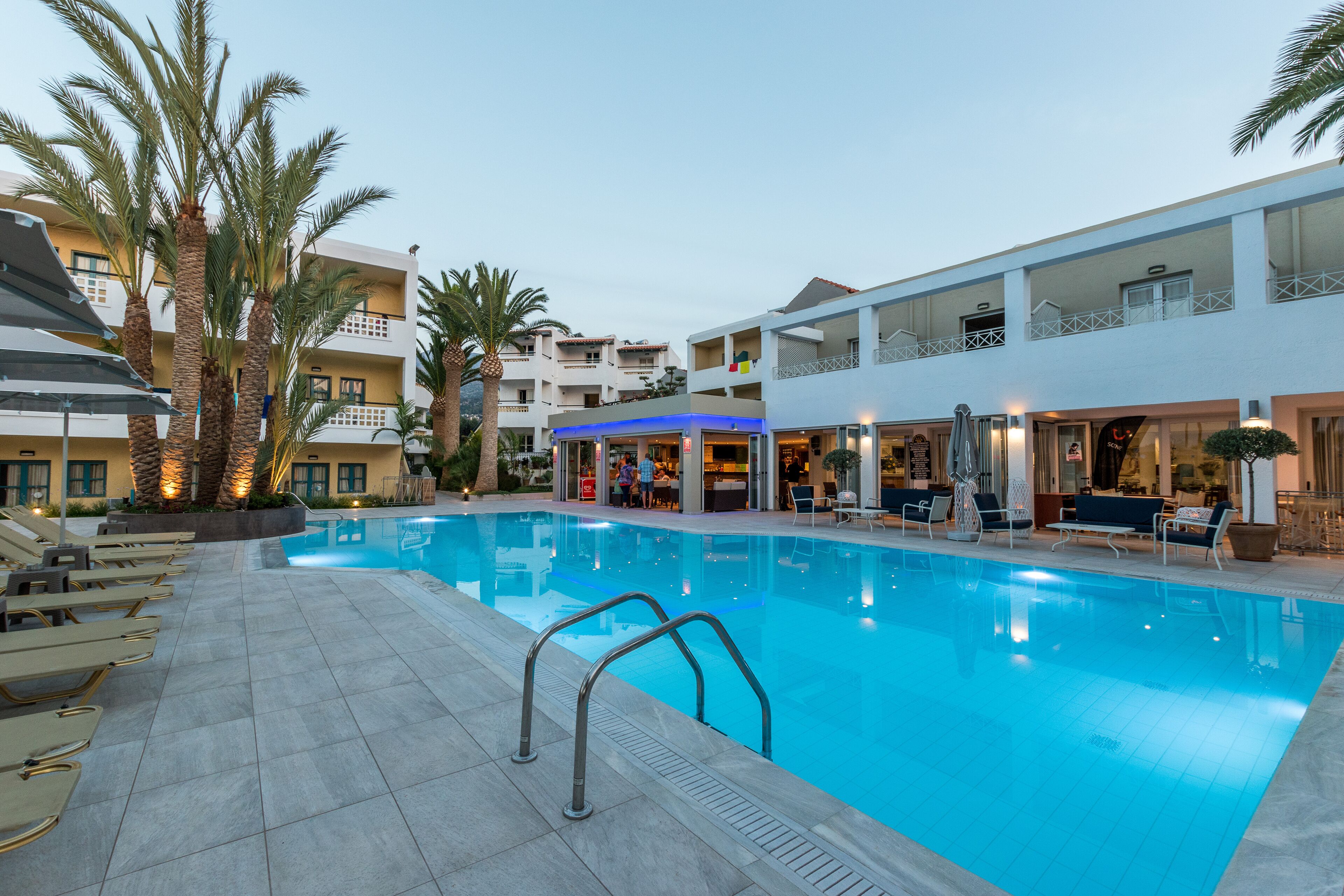 A swimming pool with lounge chairs and palm trees, surrounded by multi-story buildings with balconies.
