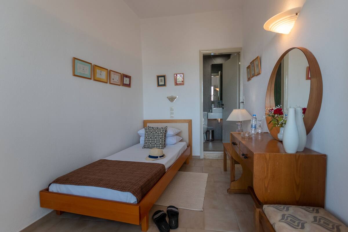 Un dormitorio con una cama individual, un aparador de madera, un espejo y una mesita de noche con un jarrón y flores.