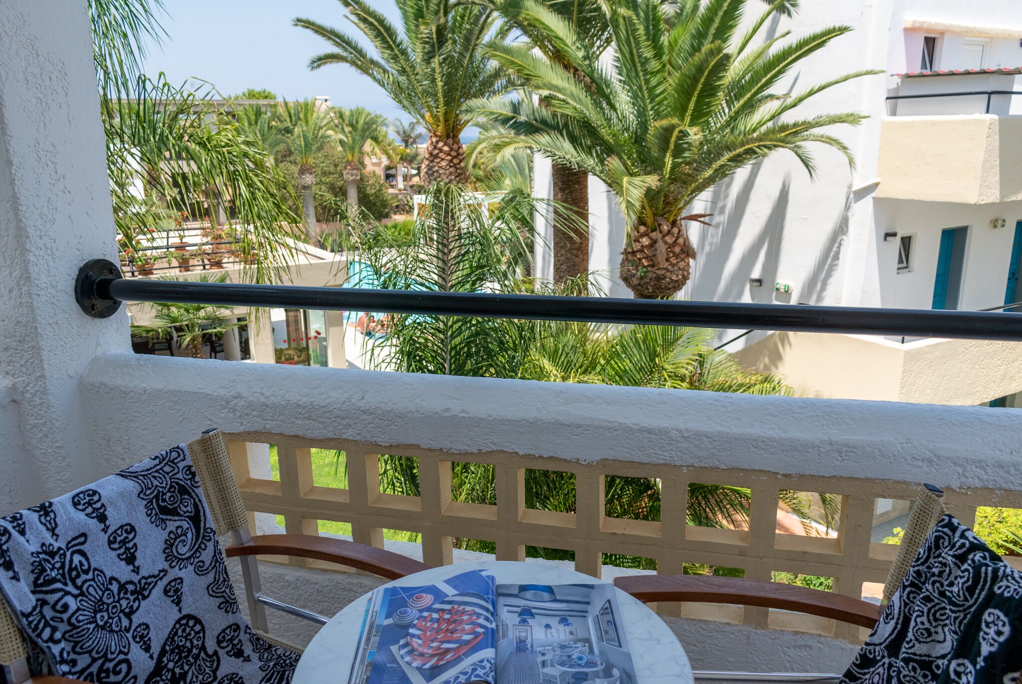 A balcony with a wooden chair and a table, overlooking a pool and palm trees.