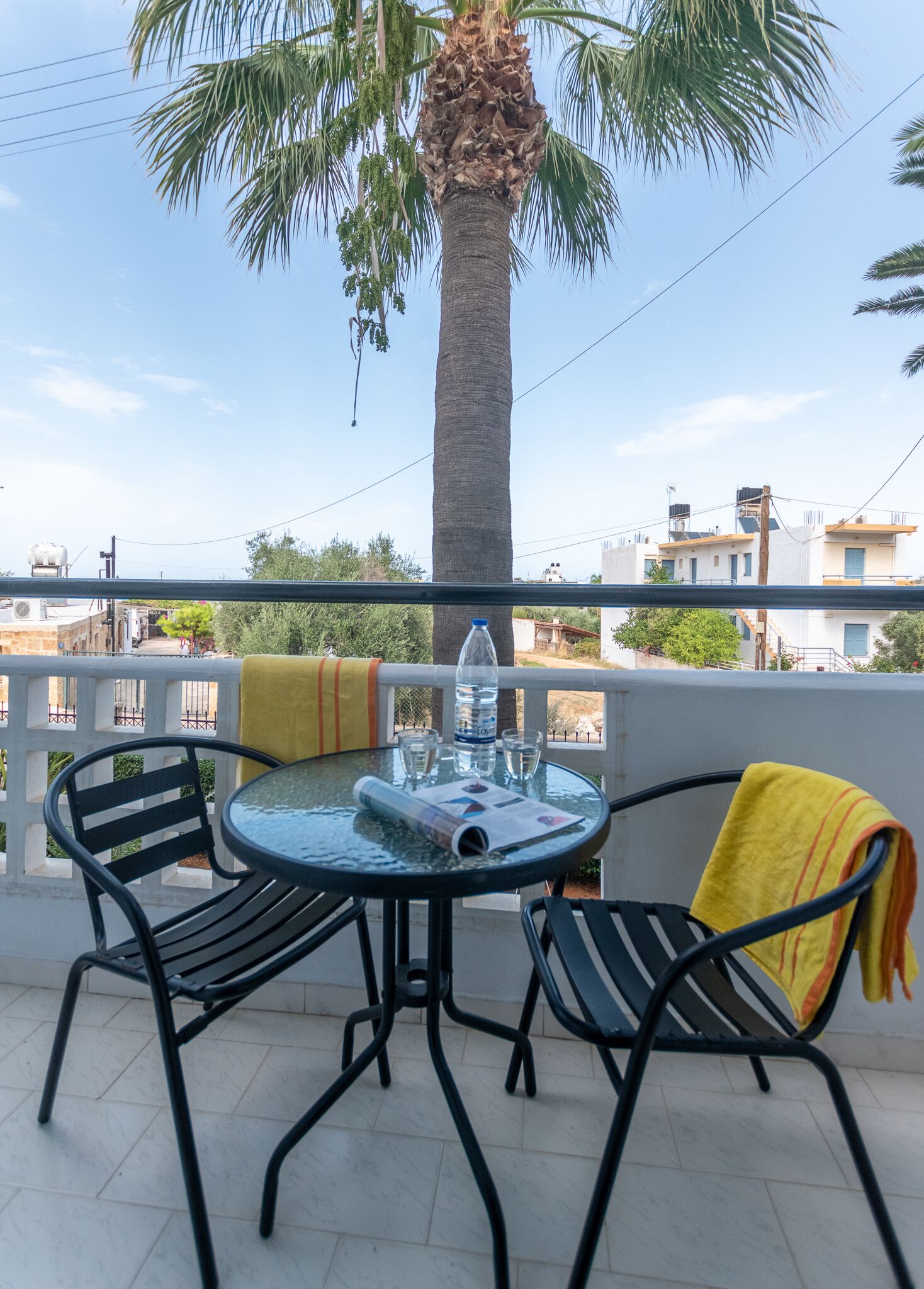 A balcony with a round glass table, two chairs, a water bottle, and a yellow towel.