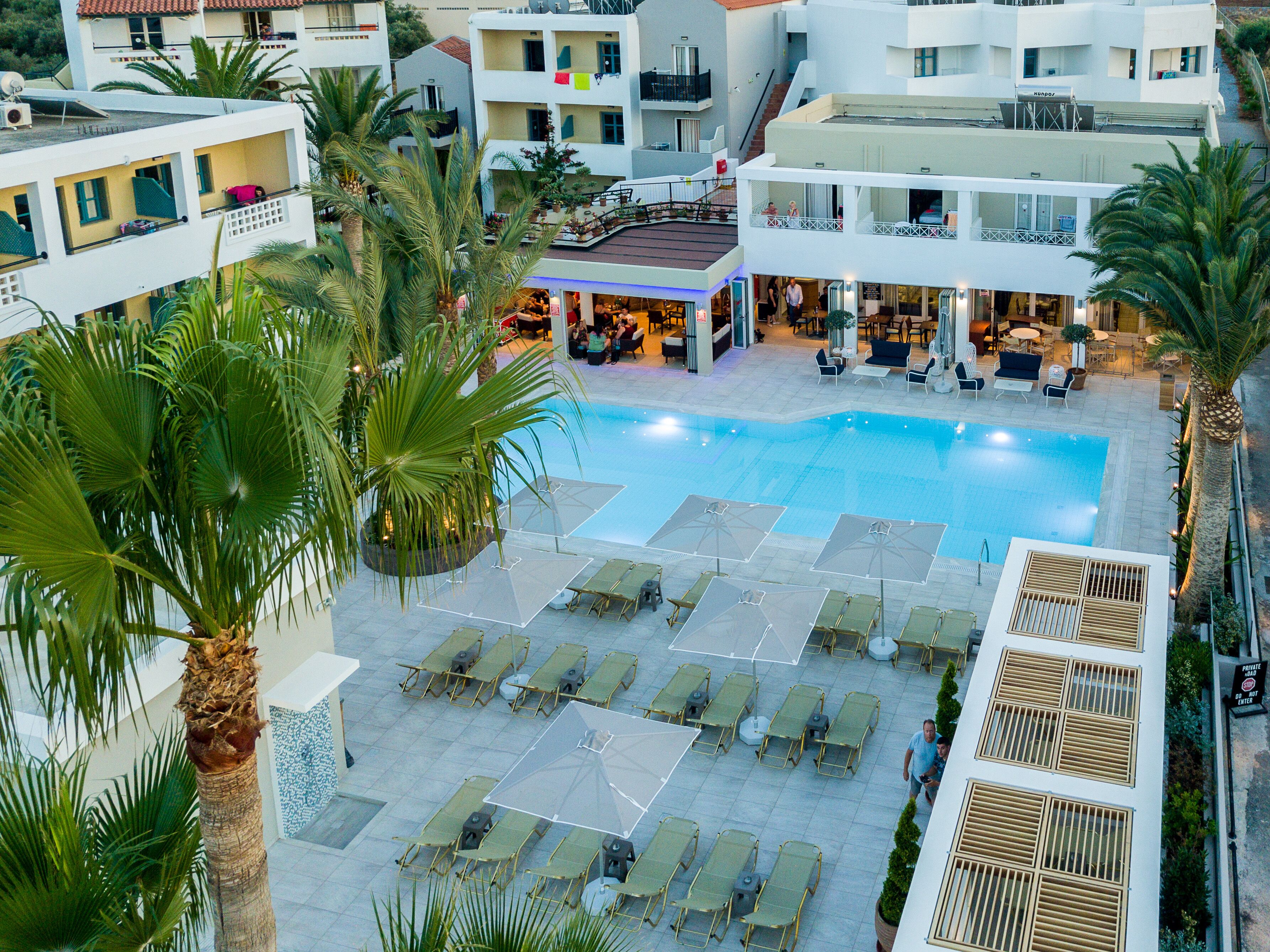 A rooftop pool area with lounge chairs and umbrellas, surrounded by palm trees and modern buildings.