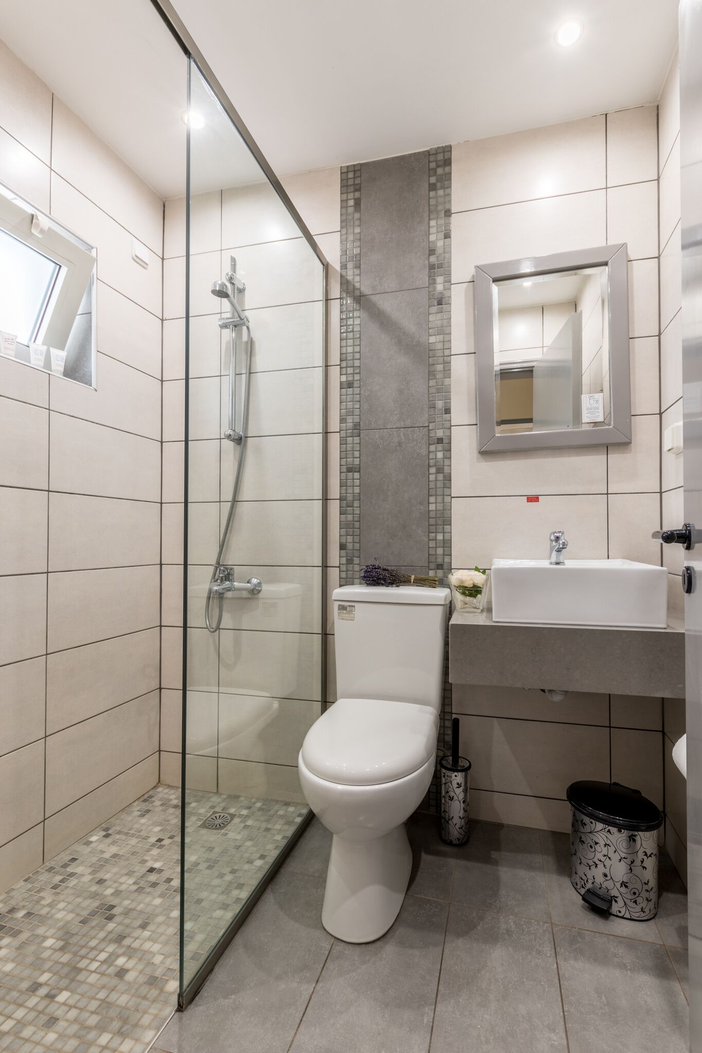 A modern bathroom with a glass-enclosed shower, a white toilet, a sink with a mirror, and a trash bin.
