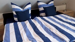 Uma cama com colcha azul e branca listrada, duas almofadas iguais, uma mesa de cabeceira com abajur e um criado-mudo de madeira.