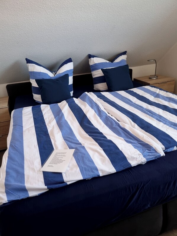 Un lit avec une couvre-lit à rayures bleues et blanches, deux oreillers assortis, une table de chevet avec une lampe et une petite table en bois.