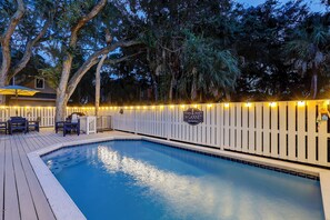 A swimming pool with a wooden deck, surrounded by a white fence with string lights.