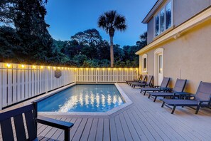 Ein Schwimmbecken mit Liegestühlen und einer Holzterrasse, umgeben von einem Zaun mit Lichterketten.